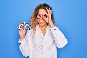 Wall Mural - Beautiful oculist woman with blue eyes holding optometry glasses over isolated background with happy face smiling doing ok sign with hand on eye looking through fingers