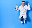 © Krakenimages.com - Middle age handsome doctor man wearing stethoscope and coat smiling happy. Jumping with smile on face holding clipboard over isolated blue background