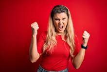 Young Beautiful Blonde Woman Wearing Casual T-shirt Standing Over Isolated Red Background Angry And Mad Raising Fists Frustrated And Furious While Shouting With Anger. Rage And Aggressive Concept.
