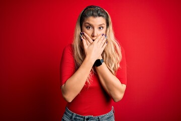 Canvas Print - Young beautiful blonde woman wearing casual t-shirt standing over isolated red background shocked covering mouth with hands for mistake. Secret concept.