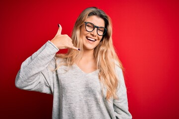 Poster - Young beautiful blonde woman wearing sweater and glasses over isolated red background smiling doing phone gesture with hand and fingers like talking on the telephone. Communicating concepts.