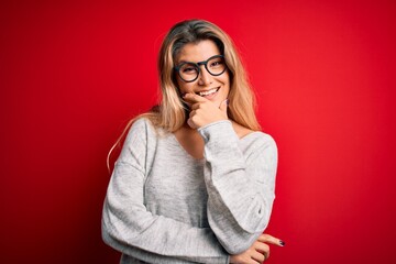 Poster - Young beautiful blonde woman wearing sweater and glasses over isolated red background looking confident at the camera smiling with crossed arms and hand raised on chin. Thinking positive.