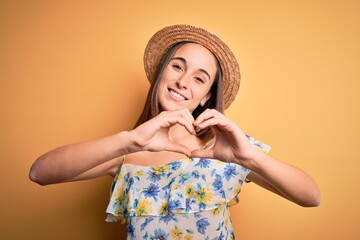 Poster - Young beautiful woman wearing casual t-shirt and summer hat over isolated yellow background smiling in love doing heart symbol shape with hands. Romantic concept.