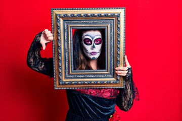 Sticker - Young woman wearing day of the dead costume holding empty frame with angry face, negative sign showing dislike with thumbs down, rejection concept