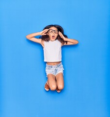 Sticker - Adorable hispanic child girl wearing casual clothes and glasses surprised with open mouth. Jumping with hands on face  over isolated blue background