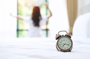 Wall Mural - Closeup alarm clock with background of happy woman stretching in bed after waking up, young adult female rising arms and looking to window in the morning. fresh relax and have a nice day concepts
