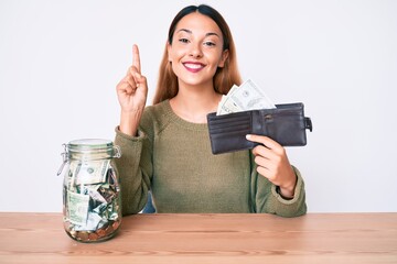 Poster - Young brunette woman with savings jar and wallet with dollars surprised with an idea or question pointing finger with happy face, number one