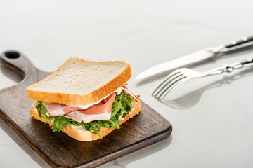 Wall Mural - selective focus of fresh sandwich with arugula and prosciutto on wooden cutting board near cutlery on white marble surface