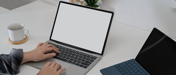 Female hands using mock-up laptop on white home office desk with digital tablet and coffee cup