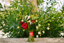 Bridal Bouquet Of Red And Pink Roses, Boxwood Branches, Not Blooming Buds Of White Flowers And Red Ribbons With Brooch Against The Background Of An Oleander Bush