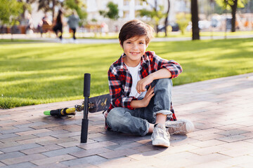 Caucasian teenager sitting on the grass in a summer sunny park with his scooter. Smiling and having fun, spending time on vacation in the city. Active lifestyle concept