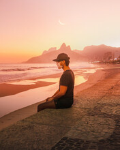 Man With A Mask Sitting On The Beach At Sunset With The Moon