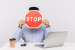 © khosrork - Man employee sitting in office workplace with laptop, hiding face behind stop symbol, warning with red traffic sign, symbol of prohibition restriction. indoor studio shot isolated on white background