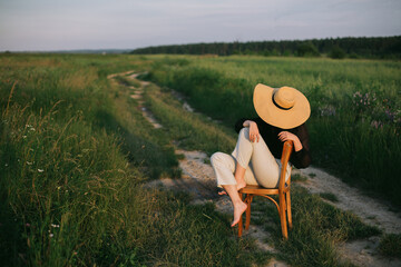 stylish elegant girl in straw hat sitting on rustic chair in summer meadow in evening. fashionable y