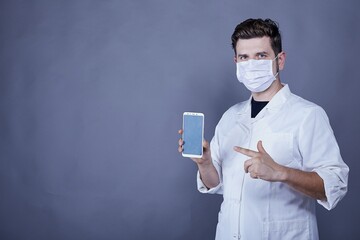 Wall Mural - Male doctor with white medical robe and a face mask showing a smartphone