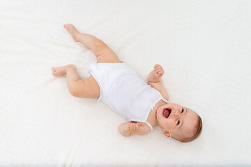 Wall Mural - baby girl 6 months old lying on the bed in the nursery on her back upside down, baby morning, baby products concept