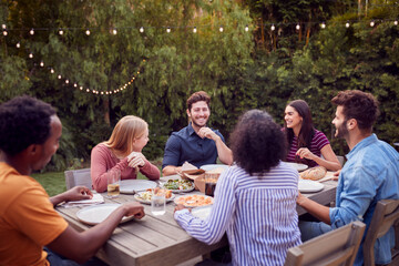 Multi-Cultural Friends At Home Sitting At Table Enjoying Food At Summer Garden Party