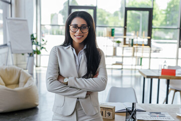 Poster - happy businesswoman in glasses standing with crossed arms near table in office