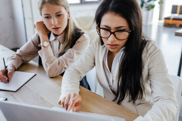 Poster - selective focus of businesswoman in glasses looking at laptop near beautiful coworker