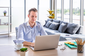 Portrait of smiling young Caucasian man freelancer working at home, sitting at desk in living room using typing laptop computer.