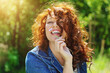 portrait jeune et jolie femme rousse aux cheveux longs bouclés et aux yeux bleus souriant dans un parc au soleil et portant une veste en jeans bleus  