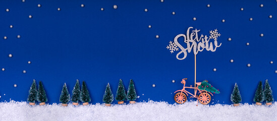 Wall Mural - Funny cozy scene with Christmas toys on classic blue. Christmas tree is moved home by bicycle on the snow road with stars shining and Let Snow wording. Horizontal banner format with place for text