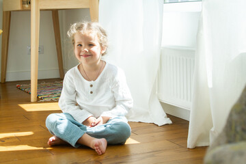 Little cute girl in a bright room, a minimum of objects in the room, the child rejoices, sitting by the window with white curtains.