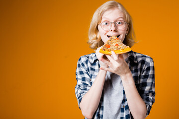 Portrait of a blonde girl in glasses who is happy to eat a slice of pizza on an orange background