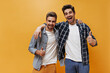 Excited brunet men in checkered shirts, colorful pants and white t-shirts smile, show thumbs up and pose on orange background.