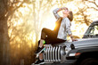 © elnariz - Solo traveler on a road trip adventure in nature, sits on hood of vehicle looking up at sky with forest sunset in the background