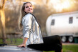 © elnariz - A beautiful smiling female traveler sitting on car in campground with RV trailer in background, sunset golden hour