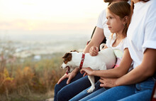 Close-up shot of the girl with her pet dog on her laps, sitting in the car trunk between her parents, family wearing white t-shirt and jeans enjoys a beautiful view from the hill