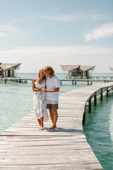 Wall Mural - Happy loving couple walking on summer tropical beach on wooden jetty with overwater wooden bungalow on background