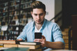 © BullRun - Concentrated millennial male checking notification on modern mobile phone while spending time in coworking space,serious young man banking via application on smartphone while learning in library .