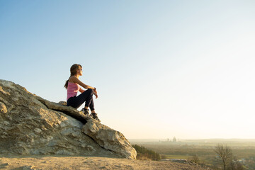 Wall Mural - Woman hiker sitting on a steep big rock enjoying warm summer day. Young female climber resting during sports activity in nature. Active recreation in nature concept.