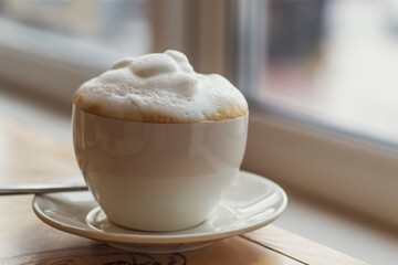 Cappuccino coffee in a white cup with lush foam on a table near the window in a cafe. Tasty breakfast.