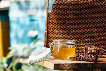 Jar of fresh honey with assorted tools for beekeeping, a wooden dispenser and tray of honeycomb from a bee hive in a still life on a wooden table outdoors 