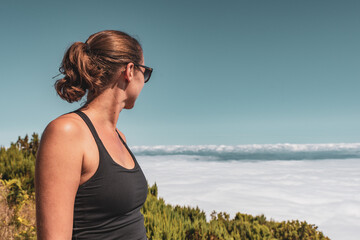 Wall Mural - Young brunette woman with sunglasses sitting at the edge of the mountain over the clouds