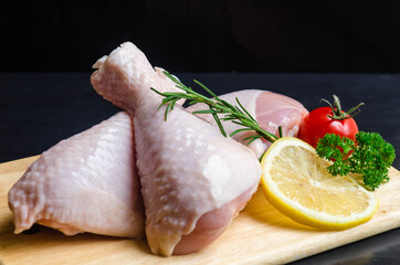 Close up of fresh rare chicken drumstick with lemon, tomato parsley and rosemary prepare for cooking on wood board and black background.