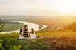 © eduard - Three female friends in elegant dresses having picnic on the hill  with river on the background. Cozy summer atmosphere. Place for text. Happy young girls friends having fun at picnic.