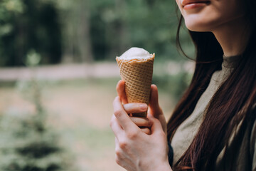 Young pretty woman holding ice cream in a waffle cone while walking along the street in the park alone.