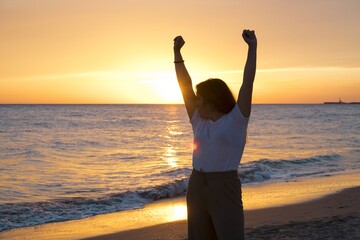 Sticker - Cheerful excited female jumping with her hands in the air on the beach at sunset