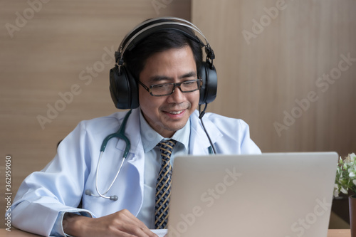 Asian man doctor wear headphones working at office desk and smiling at camera, video call consultation, patient and doctor talk concept.
