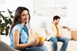 © LIGHTFIELD STUDIOS - Selective focus of woman holding cup of coffee and smiling at camera near boyfriend on sofa