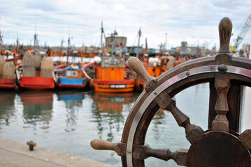 Timón de madera, con barcos naranjas de fondo estacionados en un puerto.