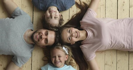 Wall Mural - Happy parents and cute little kids son daughter lying in circle on warm wooden floor, smiling dad mom and children bonding looking at camera, family portrait, underfloor heating concept, top view