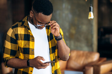 Wall Mural - Stylish African American guy, student or employee, in yellow checkered shirt standing indoor and uses his smartphone