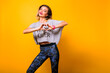 Pretty hipster woman making heart by her hands, posing at studio yellow background, wearing yoga pants and funny T-shirt.