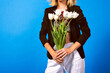 Spring studio image of woman wearing trendy suite holding bouquet of white flower tulips, toned bright colors, blue background.