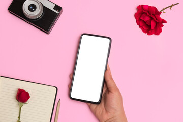 Top view of female hand holding smartphone with white blank screen on pink background with retro photo camera, notepad with pen and rose flowers. Mockup with phone